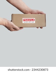 One rectangular cardboard box is held in the hands on a plain light background. A box with an adhesive warning label for packaging - Fragile, Handle with care, Thank you. No people. Corner composition