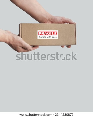 One rectangular cardboard box is held in the hands on a plain light background. A box with an adhesive warning label for packaging - Fragile, Handle with care, Thank you. No people. Corner composition