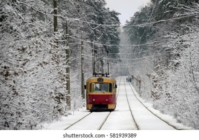 One red tram rides through the snow-covered road through the winter woods. In the distance, some people cross the road.