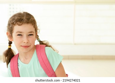 One Schoolgirl With A Backpack Stands In School Hall With Large Windows. Happy 7 Year Old Caucasian Girl. Eye Contact. Back To School, Education, Learning Concept. Indoor. Copy Space.