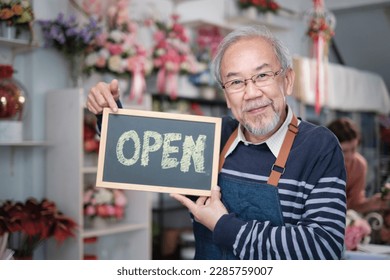 One senior male florist owner in apron shows open sign board in bright flower shop store with smile and looks at camera, small business opening, occupation retirement, happy elderly SME entrepreneur.  - Powered by Shutterstock