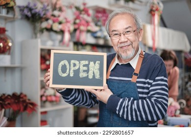 One senior male florist owner in apron shows open sign board in bright flower shop store with smile and looks at camera, small business opening, occupation retirement, happy elderly SME entrepreneur.  - Powered by Shutterstock