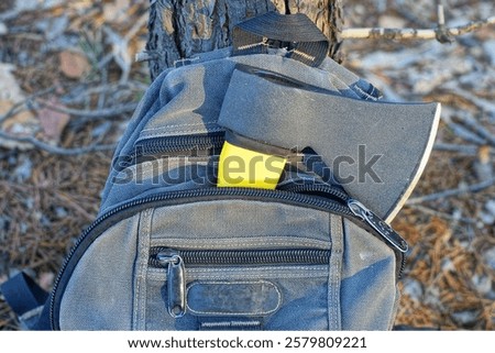 one sharp dangerous with yellow plastic handle iron modern black small hatchet in open gray with material and zipper stylish fashionable men's backpack near tree trunk daytime in forest