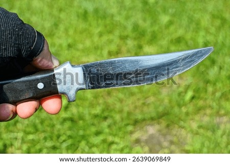 one sharp shiny gray with blade iron army with black handle dangerous industrial knife held in glove in hand by bandit on green background day outside