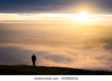 One single anonymous man standing on mountain alone watching sunrise silhouetted stood high above the clouds looking down valley with golden hour dawn sunlight on the horizon