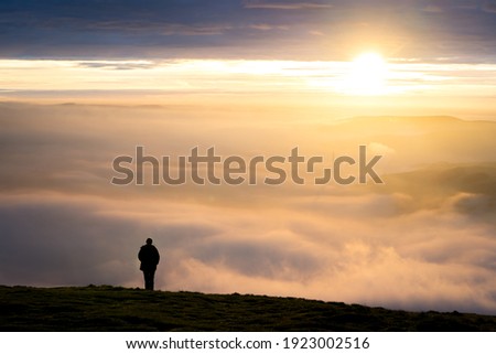 One single anonymous man standing on mountain alone watching sunrise silhouetted stood high above the clouds looking down valley with golden hour dawn sunlight on the horizon