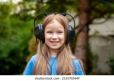 One single happy cheerful school age child, girl wearing large headphones listening to music outdoors, simple portrait, face closeup, blurred background, shallow dof, head frontal shot - Shutterstock ID 2155703449