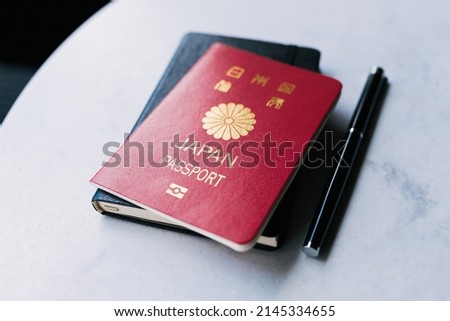 One single red Japanese passport laying on top of a black notebook next to a black pen that is on a white table