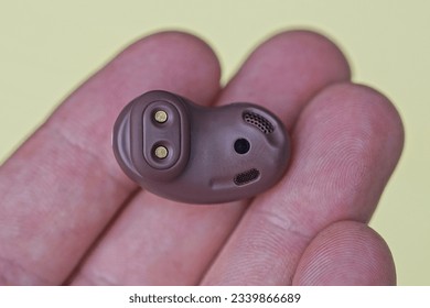 one small brown plastic wireless headphone lies on the fingers on the hand on a yellow background