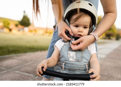 One Small Caucasian Girl Or Boy Mother Young Woman Adjust Protective Helmet Putting On Child Daughter While Playing With Kick Scooter Outdoor In Summer Day Leisure Family Growing Up Childhood Concept