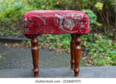 One Small Ottoman Chair With A Soft Seat Made Of Red Fabric And Brown Wooden Legs Stands On The Street Against The Backdrop Of Green Vegetation