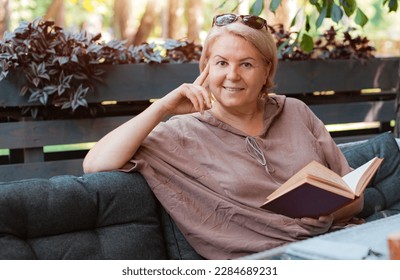One smiling elegant well-groomed elderly senior woman is reading a book in a summer cafe. Lifestyle and recreation of a retired tourist in the city. 50-60 years old age