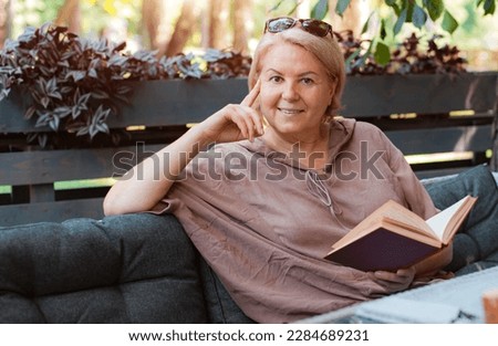 One smiling elegant well-groomed elderly senior woman is reading a book in a summer cafe. Lifestyle and recreation of a retired tourist in the city. 50-60 years old age