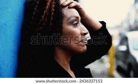 One Stressed South American black woman struggling with anxiety and worry while leaning on urban wall outside in city street. Preoccupied person suffering alone in crisis, feeling despair