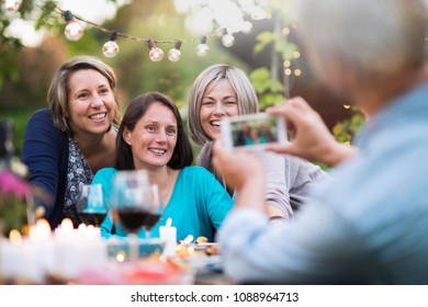 One summer evening friends gathered around a table in the garden for a good time. A man takes a picture of three female friends in their forties