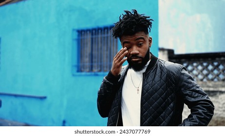 One tired young black man standing outside in city street touching face and eyes, wearing jacket in drizzle rain. 20s person after a rough night - Powered by Shutterstock