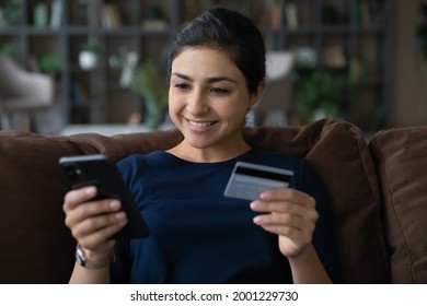 In one touch. Smiling young mixed race female client use prepaid plastic card and phone web app to book tickets online make room reservation at hotel abroad. Indian woman check ebank account by phone