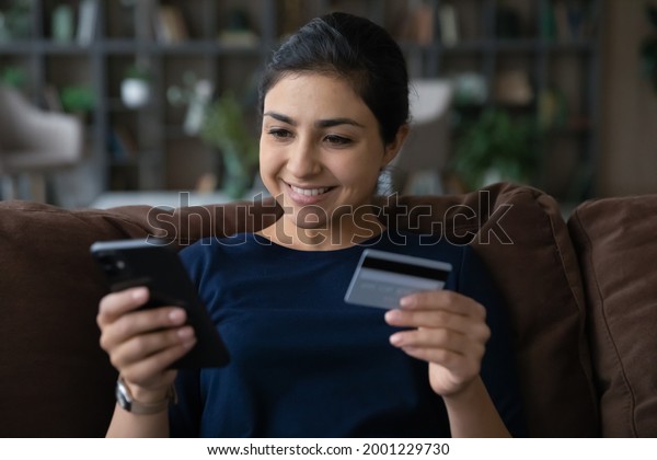 In one touch. Smiling young mixed race female
client use prepaid plastic card and phone web app to book tickets
online make room reservation at hotel abroad. Indian woman check
ebank account by phone