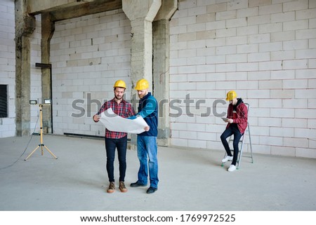 One of two engineers pointing at sketch on blueprint and specifying details while consulting with colleague on background of man with tablet