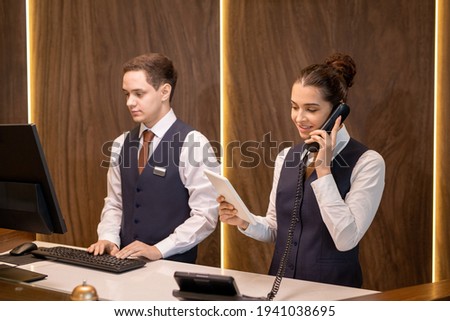 One of two young hotel receptionists standing by counter, looking at touchpad display and consulting client on the phone against colleague