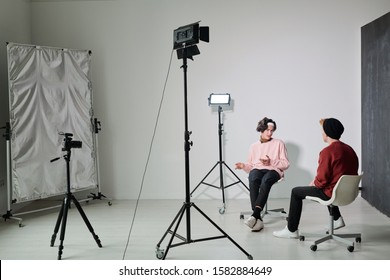 One Of Two Young Male Vloggers Explaining Something To His Friend While Both Sitting On Chairs In Front Of Each Other In Studio