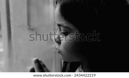One unhappy somber little girl feeling depressed during hard times. Close-up child's face and hand leaning on glass window feeling lonely and solitude in black and white, monochromatic