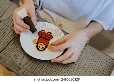 One unrecognizable Caucasian girl is sitting at a table, decorating baked cookies with chocolate from a tube, sitting at a dark gray table, close-up side view. Step by step instructions. Step 30 - Powered by Shutterstock