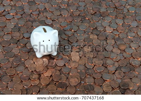 One very small white piggy bank sitting on a landscape of old and new pennies. With the collapse of the zinc market, the penny has become cheaper to make, costing the taxpayer much less.