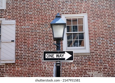 One Way Sign Hanging On A Black Pole With Street Lamp, With A Traditional Brick Wall On The Back With White Window