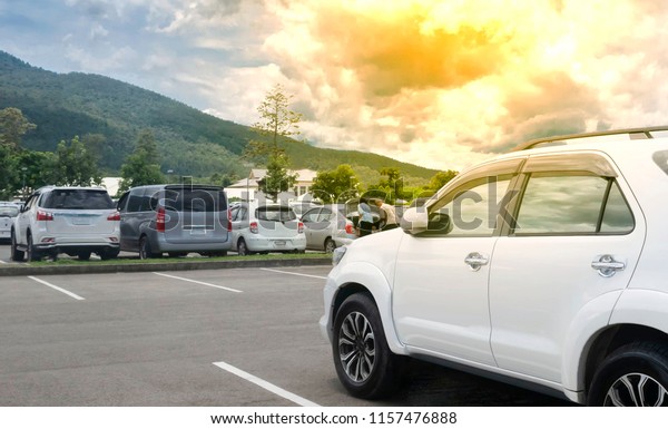 One white car parking on asphalt parking lot near\
row of others cars parking at outdoor parking lot with trees,\
mountain, beautiful sunset cloudy sky background, transportation\
with nature