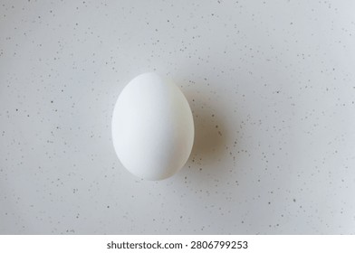 One white egg on a light background with small specks. The clean shape and smooth texture of the egg, symbolizing simplicity and naturalness. Farm to table, minimalist still life. Top view.