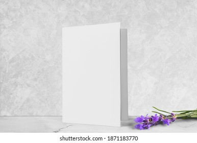 One white greeting card mockup, standing upright on a white wooden desk with flowers