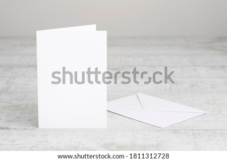 One white greeting card mockup with envelope, standing upright on a white wooden desk. Blank, closed card template. 