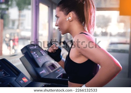 One woman with black tops and bluetooth ear phones running on a tread mill in a gym.