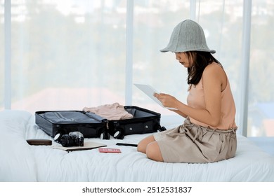 One woman in casual hat sits on bed, checking her digital tablet while packing for trip, with suitcase and travel essentials nearby.
 - Powered by Shutterstock