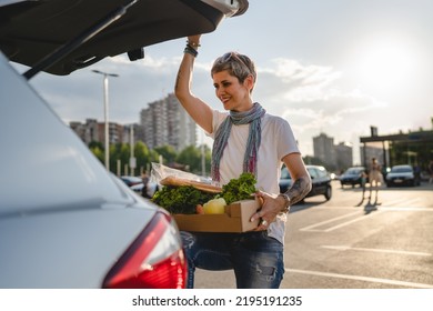 One Woman Mature Caucasian Female Standing By The Back Trunk Of Her Car On The Parking Lot Of The Supermarket Shopping Mall Or Grocery Store With Vegetables Food In Box Putting Them In The Vehicle