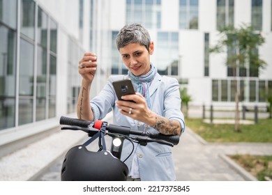 One Woman Mature Senior Caucasian Female Standing Outdoor Use Mobile Phone For Selfie Photos Or Video Call By Electric Kick Scooter In Day Confident Modern Alternative Mode Of Transport Copy Space Sms