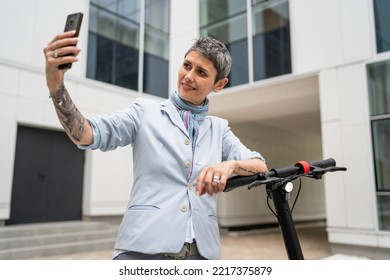 One Woman Mature Senior Caucasian Female Standing Outdoor Use Mobile Phone For Selfie Photos Or Video Call By Electric Kick Scooter In Day Confident Modern Alternative Mode Of Transport Copy Space