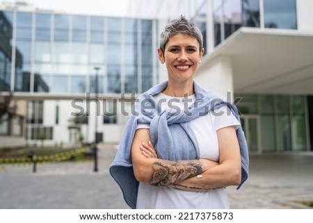 one woman mature senior caucasian female standing outdoor happy smile confident arms crossed in front of modern building in summer day short gray hair tattoo on hand copy space real people waist up