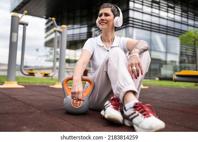 One Woman Modern Mature Caucasian Female With Short Hair Sitting In Day With Kettlebell Weight At Outdoor Gym Taking A Brake Rest After Set Workout Sport Fitness Healthy Lifestyle Concept Copy Space