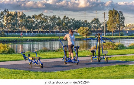 One Woman On An Exercise Machine At An Outdoor Riverbank Gym In The Early Evening.