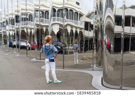 One woman standing on a street in front of a len lye (crooked) mirror and looking to her distorted reflection