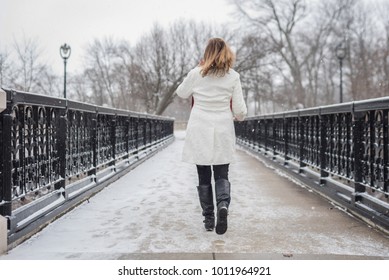 One Woman Walking On Bridge In City Park With Snow Falling And Back To Camera