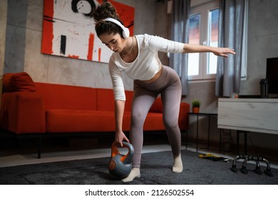 One woman young adult sporty female training at her apartment in room standing with kettlebell weight in her hands with headphones on her head full length brunette home workout side view copy space