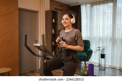 One woman young caucasian female training at home with headphones on Indoor Cycling stationary Exercise Bike real people copy space health and fitness workout hold bottle of water or supplement - Powered by Shutterstock