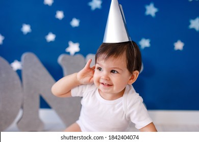 One Year Old Baby Boy In White T-shirt And Silver Hat Celebrates Birthday Near Silver Letters ONE On Purple Background With Stars.