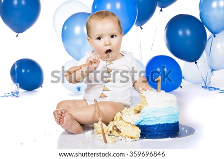 A one year old baby boy smashing a blue iced birthday cake on a silver board with lots of blue helium balloons in the background.
