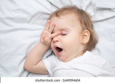 One Year Old Baby Girl Boy Wants To Sleep, Rubs His Eyes With His Hand And Yawns On White Sheets From Above. Sleep Training Mode