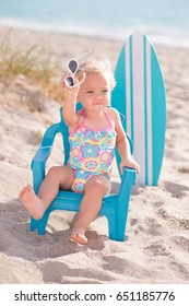 A One Year Old, Baby Girl Sitting On A Tiny Beach Chair At A Sandy Beach. She Is Holding Sunglasses And Wearing A Bathing Suit.