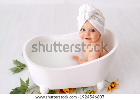 One year old baby girl takes bath. In blue swimming cap. Bathroom. The girl bathes in a basin. Clothes are dried on a hanger. Bath screens.